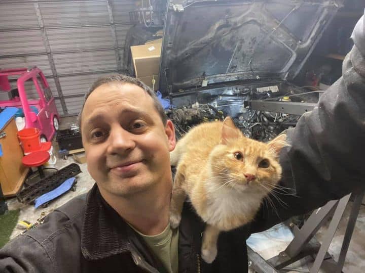 A man in a garage with a long-haired ginger cat perched on his right shoulder